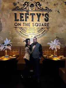 1920's speakeasy They stand amidst a bustling Great Gatsby-themed party, surrounded by fellow party-goers adorned in vintage attire, exuding an aura of glitz and glamour. The couple's radiant smiles and sophisticated poses reflect their enjoyment of the festive ambience